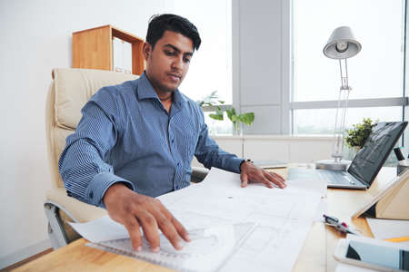 Indian architect man working with blueprints and sitting at workplace in officeの写真素材