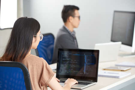 Back view of young brunette woman in glasses sitting at office desk with laptop and coding typing on keyboard on blurred backgroundの写真素材