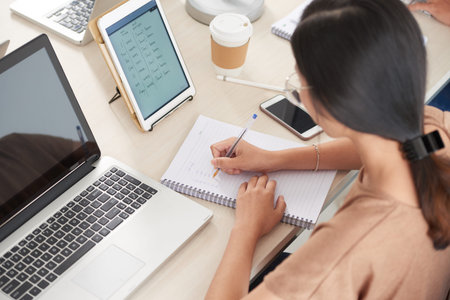 Crop back view of brunette woman in casual clothes sitting at light wooden desk with tablet and...