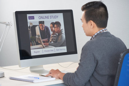 Back view of male in casual clothes sitting at computer and looking at online study site in web...