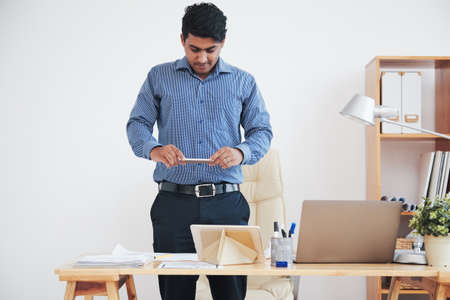 Adult Indian man taking shot of papers on desk with smartphone in officeの写真素材