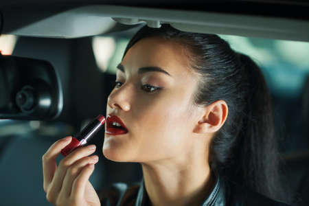 Attractive young woman looking at rearview mirror when applying bright red lipstickの写真素材