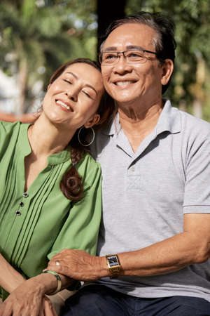Cheerful senior Vietnamese man hugging his smiling wife when they are resting in parkの写真素材