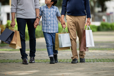 Anonymous boy holding hands of father and grandfather and waking along path after shopping on sunny...