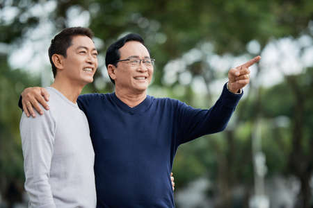 Elderly Asian man smiling and pointing at distance while embracing son on blurred background of parkの写真素材