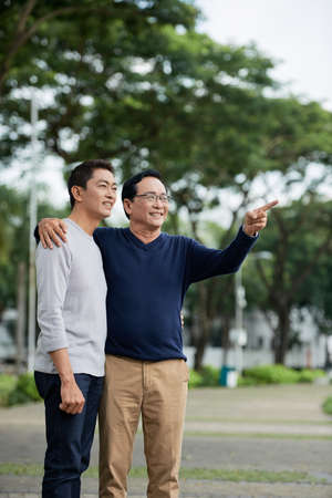 Aged Asian man embracing son and pointing at distance while standing on blurred background of parkの写真素材