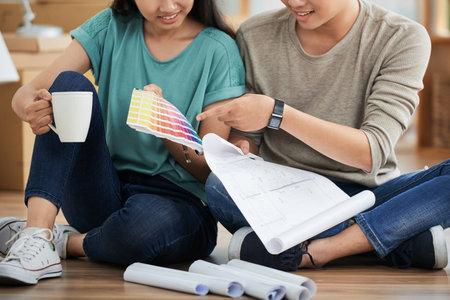 Anonymous young couple with cup of hot beverage choosing color while sitting on floor in new...