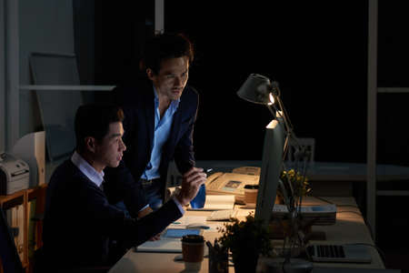 Side view of two Asian men looking at computer monitor while working late in dark office togetherの写真素材