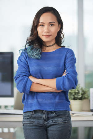 Pretty female designer keeping arms crossed and looking at camera while standing in modern officeの写真素材
