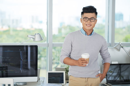 Handsome Asian guy holding cup of hot beverage and smiling while standing near desk with various...