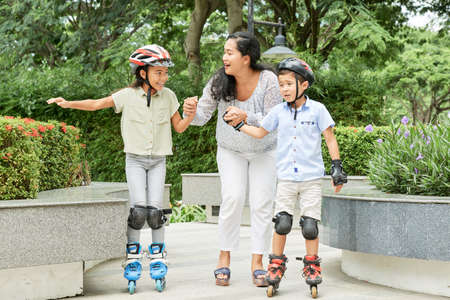 Adult Asian woman helping lovely boy and girl in helmets to ride roller skates on path in parkの写真素材