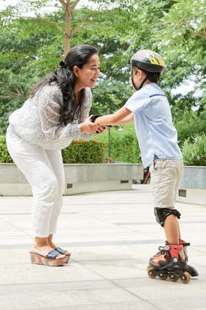 Side view of cheerful Asian woman supporting boy while teaching him to ride roller skates in parkの写真素材