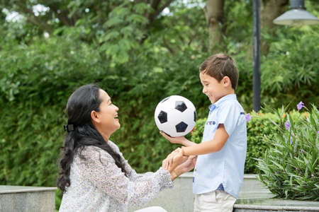Side view of cheerful Asian woman supporting cute boy with football ball while spending time in beautiful park togetherの写真素材