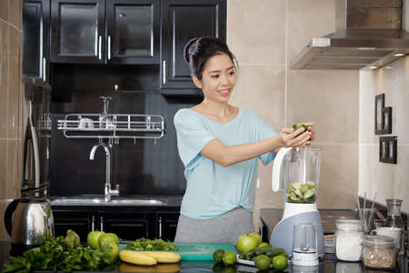 Attractive Asian female smiling and putting fresh ingredients in blender while preparing smoothie in kitchenの写真素材