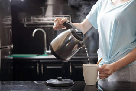 Anonymous female filling mug with hot water while brewing beverage in kitchenの写真素材