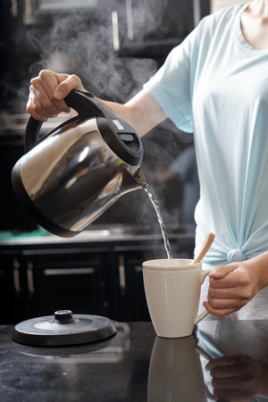 Crop woman holding kettle and filling mug with hot water while brewing drink in modern kitchenの写真素材