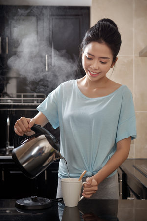 Pretty Asian lady smiling and pouring hot water into mug while brewing beverage in stylish kitchenの写真素材