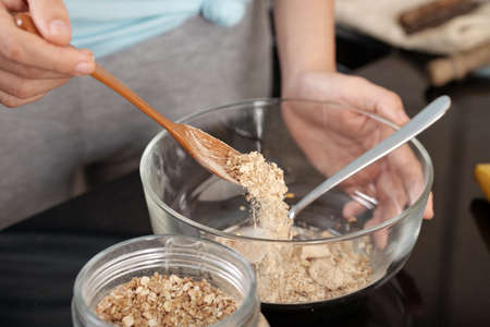 Close-up shot of crop hand using spoon to pour dry oatmeal into glass bowl while preparing breakfast in kitchenの写真素材
