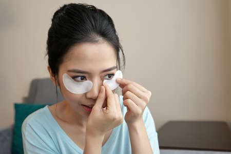 Attractive Asian woman applying under-eye patches while sitting in stylish living roomの写真素材