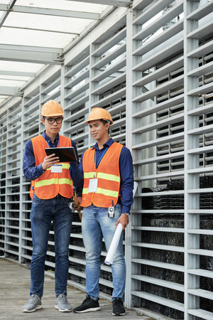 Two handsome Asian guys in hardhats and waistcoats using tablet while standing near building on...