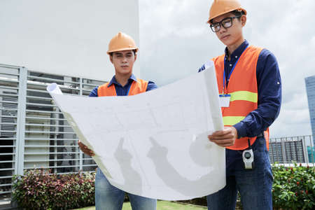 Two young Asian men in helmets and waistcoats looking at construction plan while standing on streetの写真素材