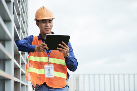 Handsome Asian man leaning on building wall and browsing tablet while standing on construction site