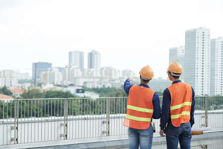 Back view of two men in helmets and waistcoats standing on construction site and looking at modern cityの写真素材