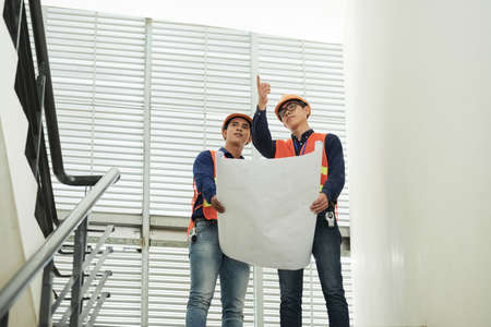 Two Asian men with draft pointing and looking at distance while standing on stairway in building under constructionの写真素材