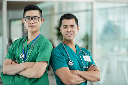 Two handsome Asian men in medical scrubs keeping arms crossed and looking at camera while standing on blurred background of hospital hallの写真素材