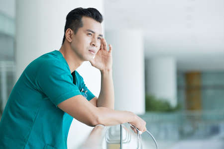 Side view of handsome Asian man in medical uniform looking away and thinking while leaning on railing in hospital hallの写真素材