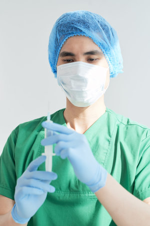 Young Asian man in medical uniform looking at syringe before injection while working in hospitalの写真素材