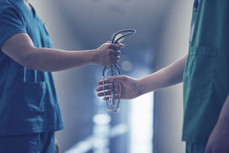 Side view of anonymous man in medical uniform passing stethoscope to male colleague while standing on blurred background of hospital corridorの写真素材
