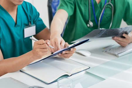 Crop health professionals with X-ray picture sitting at desk in hospital office an writing in medical recordsの写真素材