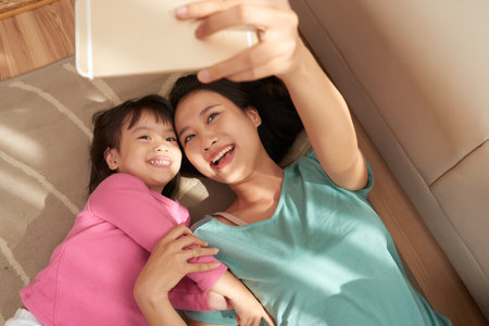 From above shot of cheerful Asian woman and girl lying on floor and taking selfie with smartphone at homeの写真素材