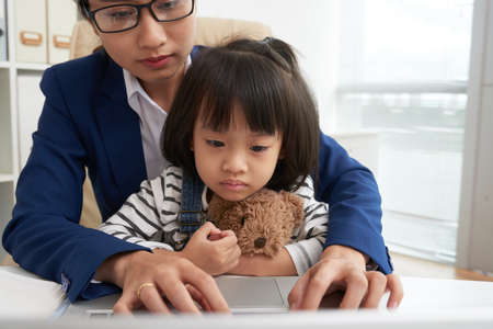 Crop Asian woman in glasses working on laptop in office with adorable little daughter on knees watching at screenの写真素材