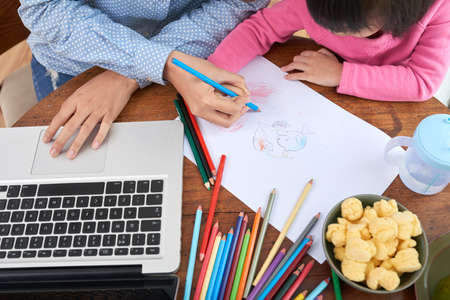 Crop shot from above of woman with laptop sitting at table with little girl helping in drawing while workingの写真素材