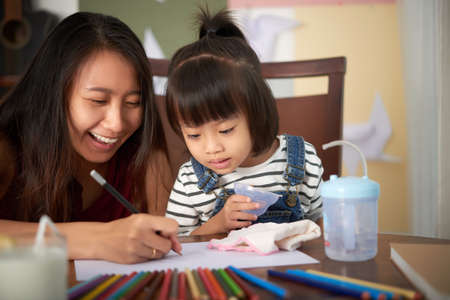 Adult Asian woman sitting at table with little daughter and drawing together on paper with colored pencils having funの写真素材