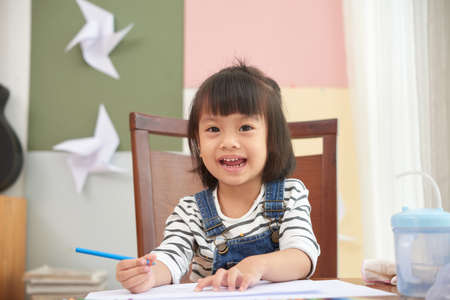 Charming Asian girl sitting at table with colored pencils looking smilingly at camera at homeの写真素材