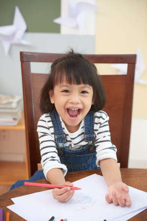 Charming Asian girl at table with paper and colorful pencils looking excitedly at cameraの写真素材