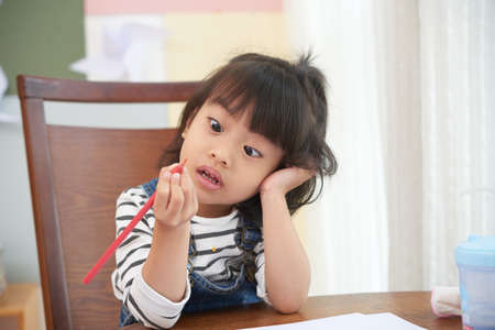 Little Asian girl sitting at table with papers and looking at colored pencil with amazement and confusionの写真素材