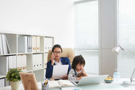 Adult businesswoman having phone call and looking papers with adorable daughter sitting on knees at workplaceの写真素材