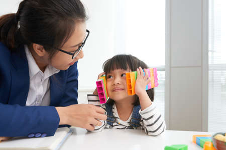 Adult businesswoman in glasses with adorable little girl sitting at table with colorful blocks inside of officeの写真素材