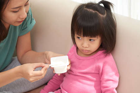 Asian woman feeding little girl sitting on sofa and giving spoon with yogurtの写真素材