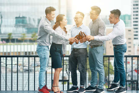 Group of happy Asian men and woman clinking with beer bottles standing outdoors on terrace with urban view on backgroundの写真素材