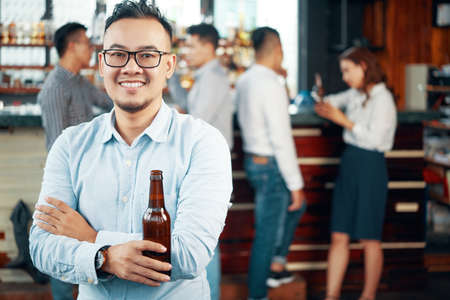 Smiling Asian man in glasses and shirt holding beer bottle and looking at camera in bar with people on backgroundの写真素材