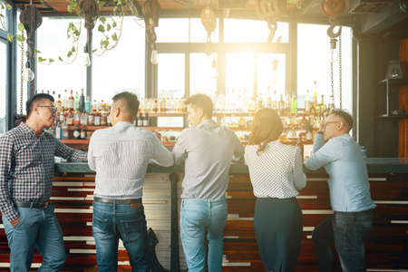 Back view of men and woman gathering at bar counter in bright sunlight having meeting togetherの写真素材