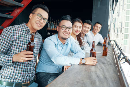 Perspective view of cheerful Asian men and woman sitting at counter near window with beer bottles and smiling at cameraの写真素材