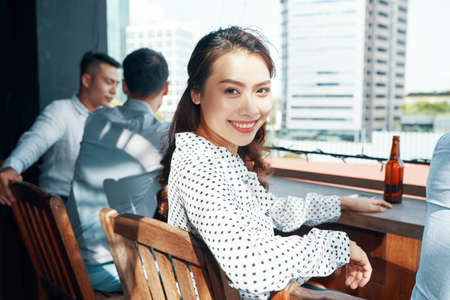 Beautiful cheerful Asian woman in polka dot shirt sitting at wood counter with beer bottle smiling at cameraの写真素材