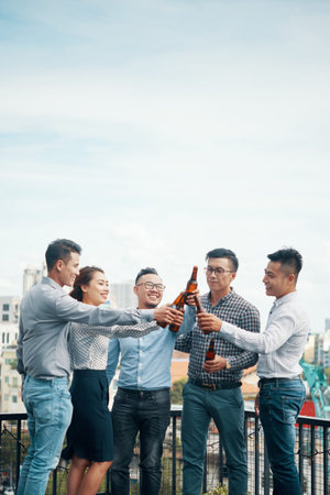 Smiling Asian men and woman standing in group on urban terrace and clinking with beer glass bottles