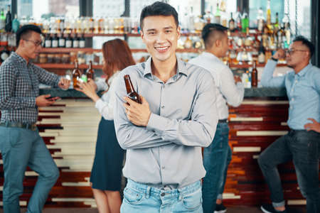Handsome Asian man in shirt holding glass bottle of beer and smiling at camera in bar with colleaguesの写真素材